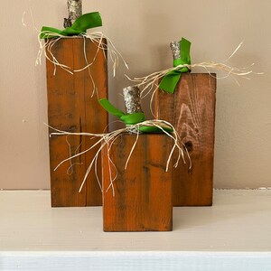 May include: Three wooden pumpkins decorated with green ribbon and straw. The pumpkins are different sizes and are arranged in a row on a white surface.