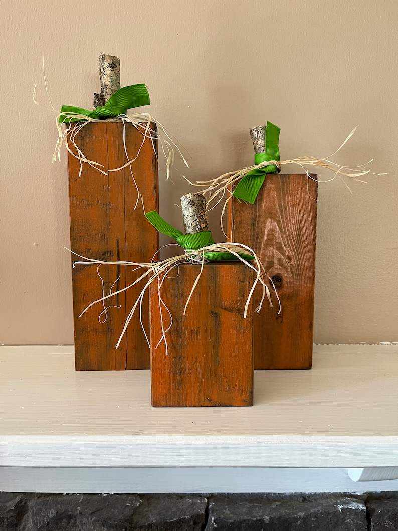 May include: Three wooden pumpkins decorated with green ribbon and straw. The pumpkins are different sizes and are arranged in a row on a white surface.