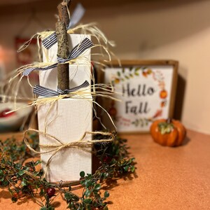 May include: A white wooden tiered stand decorated with twine, black and white checkered ribbon, and a natural twig. The stand is surrounded by a green garland with red berries and is sitting on a brown surface. A sign that says "Hello Fall" is visible in the background.