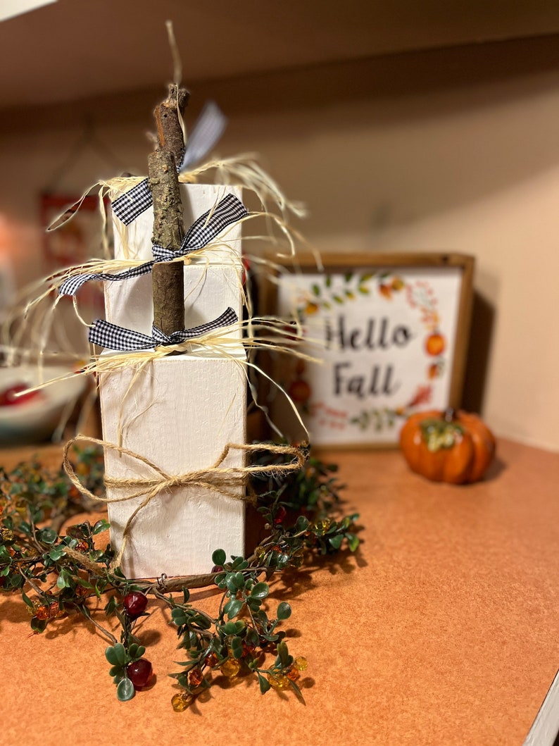 May include: A white wooden tiered stand decorated with twine, black and white checkered ribbon, and a natural twig. The stand is surrounded by a green garland with red berries and is sitting on a brown surface. A sign that says "Hello Fall" is visible in the background.
