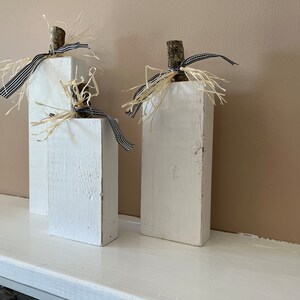May include: Three white wooden pumpkins with a rustic finish, decorated with burlap and black and white checkered ribbon.