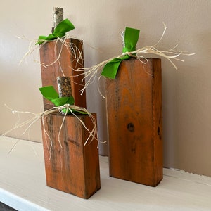 May include: Three wooden pumpkins decorated with green ribbon and raffia. The pumpkins are painted a rustic orange color and are arranged on a white shelf.