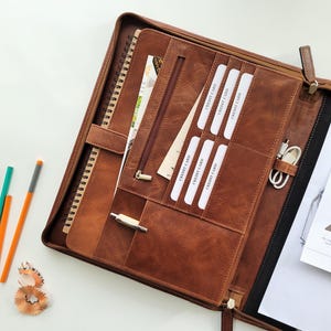 May include: A brown leather portfolio with a zippered pocket, card slots, and a pen holder. The portfolio has a built-in notepad and a clasp to keep it closed.