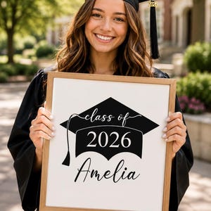 Könnte beinhalten: Ein rechteckiger Holzrahmen mit einem weißen Schild, das eine schwarze Doktorhut-Grafik, den Text "class of 2026" in einer Mischung aus Schreib- und Druckschrift sowie den Namen "Amelia" in schwarzer Schreibschrift unten zeigt.