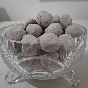 May include: A clear glass bowl, decorated with cut-glass patterns, filled with numerous round, dusty-purple sweets. The bowl rests on three small feet. The sweets are coated in a fine powder, giving them a textured appearance.