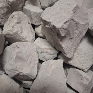 May include: A collection of light grey clay chunks, each with a rough, uneven surface. The pieces are of varying sizes and shapes, creating a textured appearance. The image is well-lit, showing the natural colour and texture of the clay.