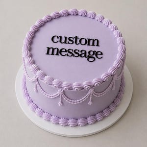 May include: A round lavender cake with smooth icing, featuring a piped border of rosettes along the top and bottom edges. The sides are decorated with delicate piped swags and beads. The text custom message is centred on top in black.