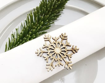 May include: A festive table setting with a white napkin held by a wooden snowflake ring. A sprig of green pine and silverware are arranged on a gray plate, creating a holiday theme.