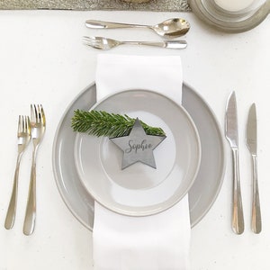 May include: A festive table setting with a gray plate, white napkin, and silverware. A star-shaped place card with the name "Sophie" is on the plate, adorned with a sprig of greenery. A lit candle and a glass cloche add to the elegant decor.
