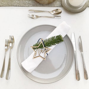 May include: A table setting with a gray plate, white napkin, and wooden star-shaped place card that reads "Mark". Silverware and a sprig of greenery adorn the napkin. A lit candle sits on a wooden base.
