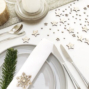 May include: A festive table setting with gray plates, silverware, and a white napkin adorned with a wooden snowflake. Scattered wooden star-shaped ornaments with words like "Joy" and "Noel" add a holiday touch. A sprig of evergreen adds a natural element.