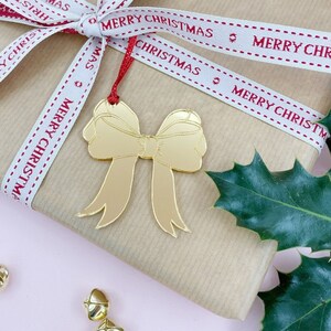 Może przedstawiać: Prezent świąteczny zapakowany w brązowy papier, przewiązany wstążką z czerwonym napisem "MERRY CHRISTMAS". Z wstążki zwisa złota zawieszka w kształcie kokardy. Liście ostrokrzewu i złote dzwoneczki zdobią różowe tło.