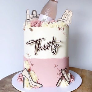 Op de afbeelding: Een verjaardagstaart met twee lagen met witte en roze glazuur. De taart is versierd met een champagnefles, hoge hakken, lippenstift en een handtas. Het woord "Thirty" staat op de voorkant.