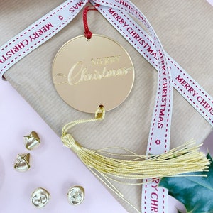 Może przedstawiać: Zapakowany prezent świąteczny ze złotą etykietą "Merry Christmas" i złotym frędzlem. Prezent jest zapakowany w brązowy papier i zawiązany białą wstążką z napisem "MERRY CHRISTMAS" w kolorze czerwonym. Złote dzwonki są rozrzucone wokół prezentu.