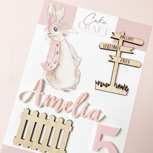 May include: A cake decoration set with a rabbit illustration, a wooden signpost, a wooden fence, the name "Amelia" in pink script, and a pink number 5. The signpost reads "Amelia's Vegetable Patch". The set is for a child's birthday.