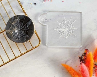 May include: A Halloween-themed image featuring a cookie and a stencil. The cookie is round with a black and gold web design and a small spider. A clear plastic stencil with a spider web design is next to the cookie. The cookie is on a gold wire rack.