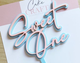 Puede incluir: Un adorno para tarta en capas con las palabras "Sweet One" en fuente cursiva. El texto es rosa claro, delineado en azul claro y sobre un fondo azul claro. El adorno tiene un tallo azul claro y el texto "Cake Craft" es visible.