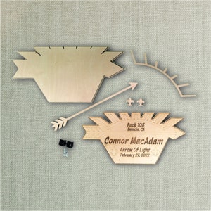 May include: Wooden craft kit for an Arrow of Light ceremony. The kit includes a wooden plaque with the text "Pack 108 Benicia, CA Connor MacAdam Arrow of Light February 27, 2022", a wooden arrow, a wooden sunburst, and two small wooden fleur-de-lis.