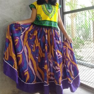 Puede incluir: Un conjunto de dos piezas que consta de una blusa amarilla brillante de manga corta con un cuello estampado en verde y negro, y una falda larga y fluida con un estampado abstracto de remolinos en tonos morado, dorado y blanco con dobladillo morado.