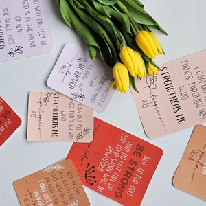 Puede incluir: Un conjunto de tarjetas inspiradoras con detalles florales y letras manuscritas. Las tarjetas presentan versículos bíblicos como "Todo lo puedo en Cristo que me fortalece" y "Sé fuerte". Tulipanes amarillos y hojas verdes están dispuestas alrededor de las tarjetas.