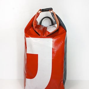 Könnte beinhalten: Eine rot-weiße Tragetasche mit einem großen weißen "J" auf der Vorderseite. Die Tasche hat einen schwarzen Griff und einen schwarzen und orangefarbenen Riemenverschluss. Die Tasche besteht aus einem glänzenden, wasserdichten Material.