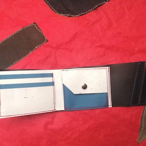 Op de afbeelding: Een drievoudig gevouwen portemonnee in wit en blauw met een zwarte stoffen achterkant. De portemonnee heeft een drukknopsluiting en een klein vakje aan de voorkant.