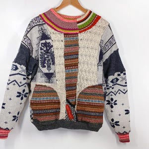 May include: A patchwork sweater with a variety of patterns and textures. The sweater features a mix of colors, including gray, blue, white, brown, and red. The sleeves have a snowflake design, and the cuffs are red. The sweater is hanging on a wooden hanger.