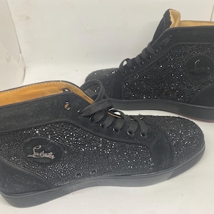 May include: A pair of black high-top sneakers with a black suede upper covered in rhinestones. The sneakers have a red rubber sole and a white logo that says "Christian Louboutin".