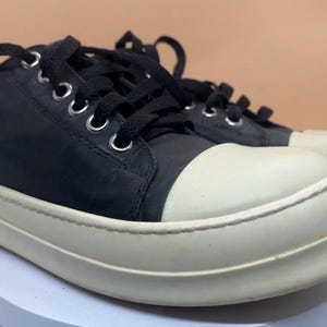 May include: A pair of black and off-white sneakers. The sneakers have black laces, a black upper, and a thick off-white sole. The toe cap is also off-white. The shoes are on a white surface.