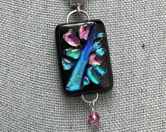 Könnte beinhalten: Ein schwarzer Glasanhänger mit schillernden blauen, grünen und rosa Akzenten. Der Anhänger ist an einer silbernen Kette befestigt und hat einen kleinen rosa Kristall, der unten herunterhängt.
