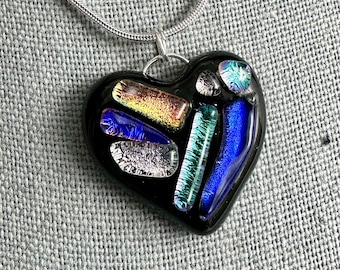 Könnte beinhalten: Eine Halskette mit einem schwarzen herzförmigen Glasanhänger mit bunten Glasstücken im Inneren. Die Kette ist aus einer silbernen Kette gefertigt.