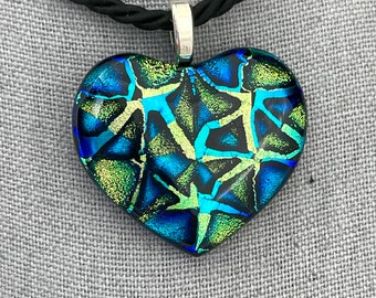 Könnte beinhalten: Eine Halskette mit einem herzförmigen Glasanhänger an einem schwarzen Band. Der Anhänger zeigt ein Mosaikdesign aus blauem, grünem und goldenem Glas.