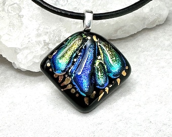 Könnte beinhalten: Ein schwarzer Glasanhänger mit einem blauen, grünen und goldenen irisierenden Design. Der Anhänger ist an einer schwarzen Lederkordel befestigt.