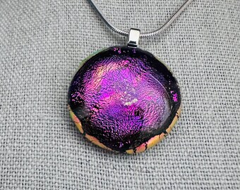 Könnte beinhalten: Eine silberne Kettenkette mit einem runden, schillernden Glasanhänger. Der Anhänger hat eine violette, rosafarbene und schwarze Farbgebung mit einer strukturierten Oberfläche.