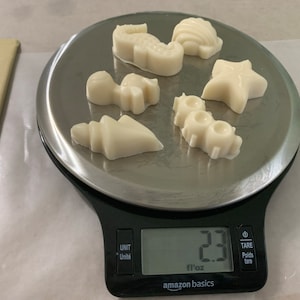 May include: A collection of white, sea-themed wax melts, including a seahorse, starfish, and seashell, are arranged on a silver scale. The scale reads 2.3 fl oz. The scale is black and has the Amazon Basics logo.