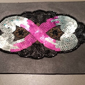 May include: A black fabric patch with a sequined infinity symbol design. The infinity symbol is made of silver, white, and pink sequins.