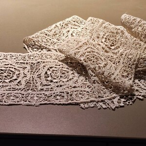 Könnte beinhalten: Eine zarte weiße Spitzenborte mit einem sich wiederholenden Muster aus filigranen Blumendesigns. Die Spitze ist auf einer braunen Oberfläche gefaltet und drapiert.