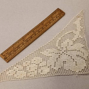 May include: A white crocheted lace triangle with a floral design. The lace is delicate and intricate, with a fine mesh pattern. The triangle is approximately 15 inches wide and 15 inches tall.