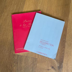 Könnte beinhalten: Zwei Auktionskataloge. Einer ist leuchtend rosa mit dem Text "Paris à la Mode HAUTE COUTURE" und dem Sotheby's-Logo. Der andere ist hellblau mit dem Text "RESURRECTION Avant-Garde Fashion" und dem Christie's-Logo.