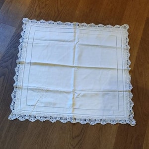 Könnte beinhalten: Weiße quadratische Tischdecke mit einer zarten Spitzenbordüre. Die Tischdecke hat ein dezentes Stickmuster in der Mitte.