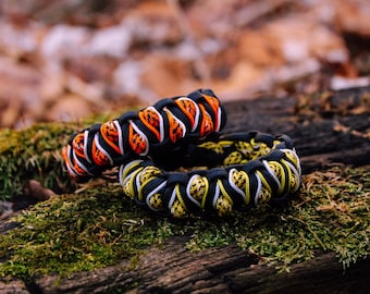 Può includere: Due braccialetti in paracord neri con strisce bianche e colorate. Il braccialetto in alto ha strisce arancioni e bianche, e il braccialetto in basso ha strisce gialle e bianche. I braccialetti sono su un tronco ricoperto di muschio.