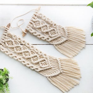 May include: Two macrame wall hangings with a natural, cream color and fringe. The hangings are made with cotton cord and are attached to wooden dowels.