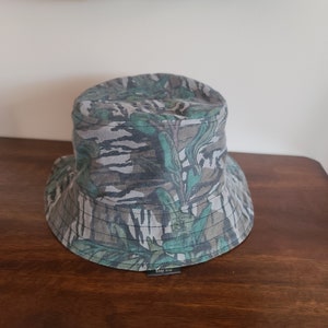 May include: A green and brown camouflage bucket hat with a black label that says "Etsy Shop".
