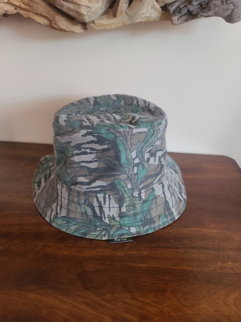 May include: A green and brown camouflage bucket hat with a black label that says "Etsy Shop".