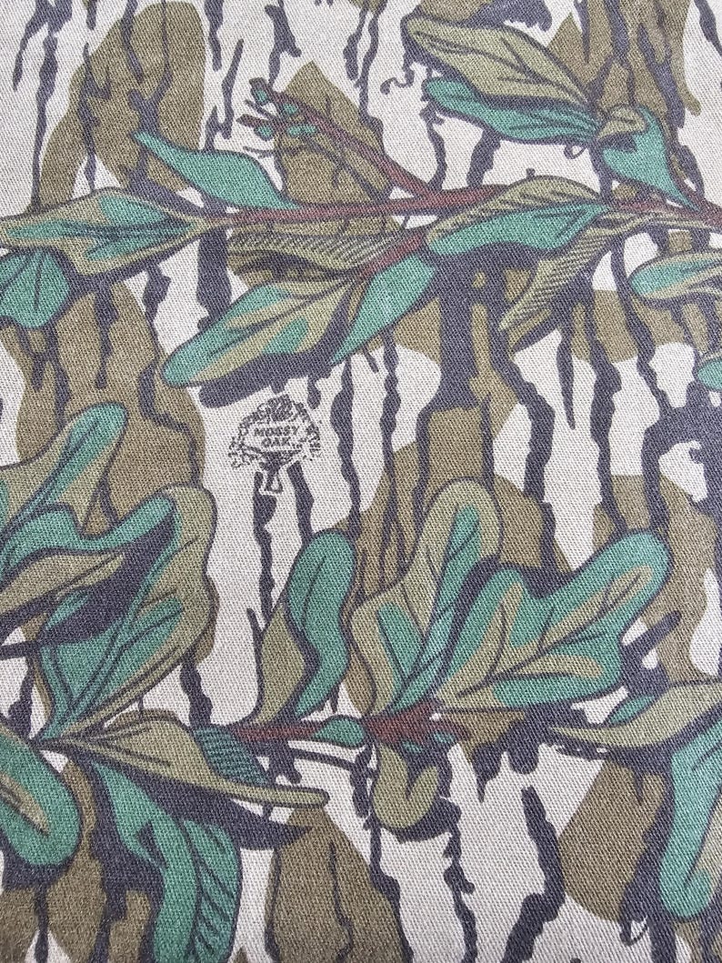 May include: Camouflage fabric with a green, brown and white pattern. The fabric features a repeating design of leaves and branches. The fabric is made from a soft, durable material.
