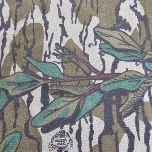 May include: Close-up of a camouflage fabric with a brown, green, and white pattern. The fabric has a textured surface and a small, circular logo that says "Mossy Oak" in white text.