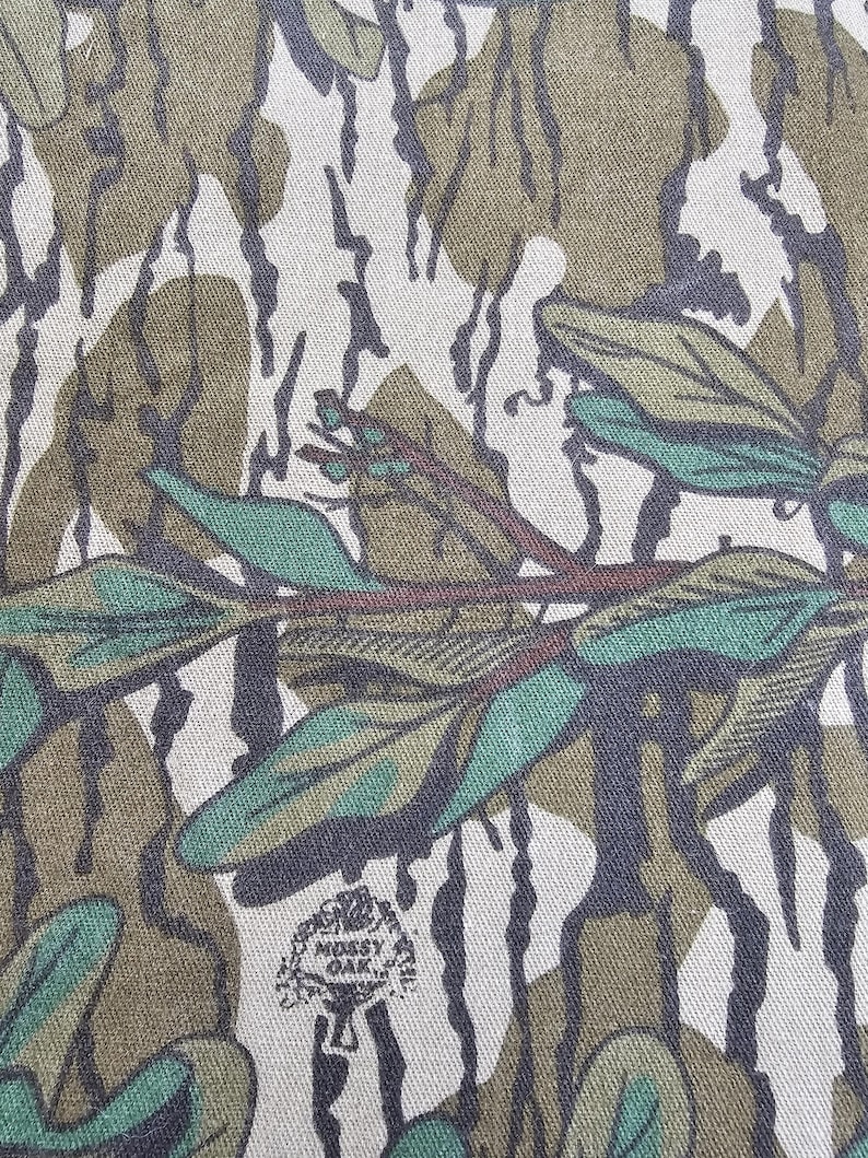 May include: Close-up of a camouflage fabric with a brown, green, and white pattern. The fabric has a textured surface and a small, circular logo that says "Mossy Oak" in white text.