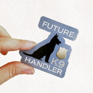May include: A blue and white sticker with the text "FUTURE HANDLER K9" and a silhouette of a German Shepherd dog. The dog is facing left and has its mouth open.