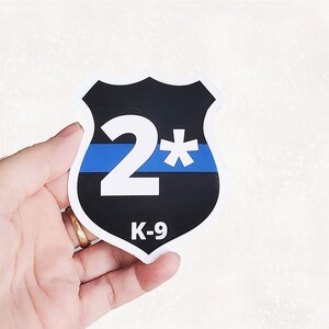 May include: A shield-shaped sticker with a black background, a blue line, and the text "2*" in white. The bottom of the sticker reads "K-9" in white. The sticker is held in a hand.