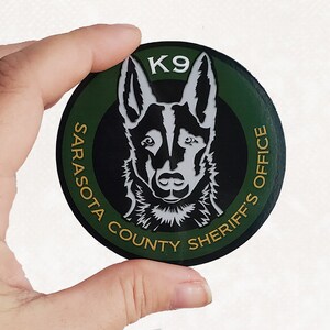 May include: A round patch featuring a K9 dog illustration. The patch has a green background with the text "SARASOTA COUNTY SHERIFF'S OFFICE" in gold lettering. The dog illustration is in black and white, with the word "K9" above it.